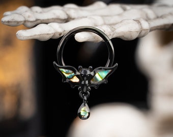May include: Black body jewelry featuring a bat charm with iridescent wings and a teardrop dangle. The bat is made of metal and has a black finish. The teardrop dangle is also made of metal and has a black finish.