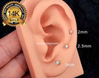 May include: Three gold ear studs with clear gemstones, shown on a model ear. The studs are 2mm, 2.5mm, and 3mm in size.  The image also includes a logo for 14K gold.