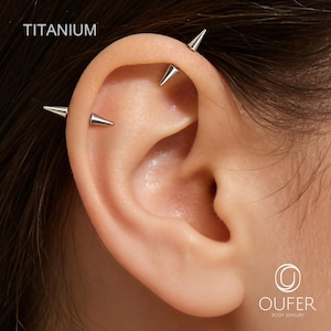 May include: Close-up of an ear with multiple silver titanium industrial piercings. The piercings are in the shape of small cones. The word "TITANIUM" is printed in white at the top left of the image. The logo for Oufer Body Jewelry is in the bottom right.