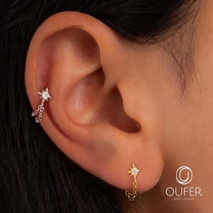 Puede incluir: Primer plano de una oreja adornada con dos pendientes en forma de estrella. Uno es plateado con cadenas colgantes, y el otro es dorado con una cadena. Ambos pendientes tienen una gema central. La imagen muestra joyería corporal.