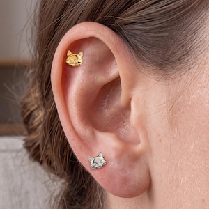 May include: Two cat-shaped stud earrings are worn in the ear. The upper earring is gold-toned with a smooth, polished finish. The lower earring is silver-toned and features small, clear, sparkling stones set into the cat's eyes and face.