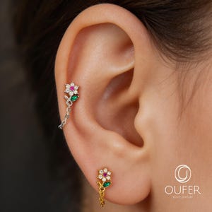 Puede incluir: Un par de pendientes con forma de flor. Un pendiente está sujeto a la oreja con una cadena y el otro es un perno. Cada flor tiene piedras preciosas blancas, rosas y verdes. Los pendientes son de color oro y plata.