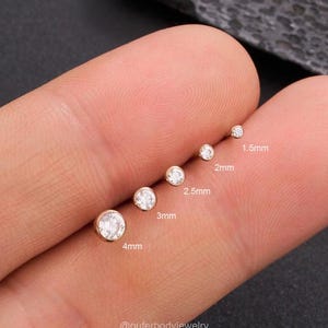 Può includere: Una collezione di orecchini a bottone color oro con gemme rotonde e trasparenti. Gli orecchini variano in dimensioni, da 1,5 mm a 4 mm. L'immagine include il testo che indica la dimensione di ogni orecchino. È visibile anche il nome del marchio "@outerbodyjewelry".