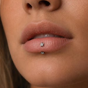 May include: A close-up of a person's lips featuring a star-shaped lip piercing. The silver-coloured piercing is on the lower lip. The skin tone is light with a soft focus.
