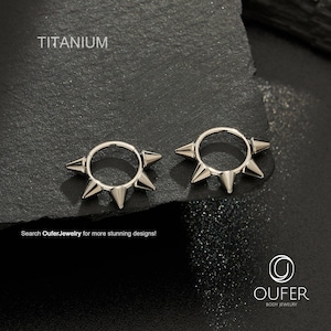 Op de afbeelding: Twee zilverkleurige titanium oorringen met een reeks scherpe, conische stekels die naar buiten steken vanaf de cirkelvormige band. De tekst TITANIUM is zichtbaar linksboven en OUFER BODY JEWELRY rechtsonder.