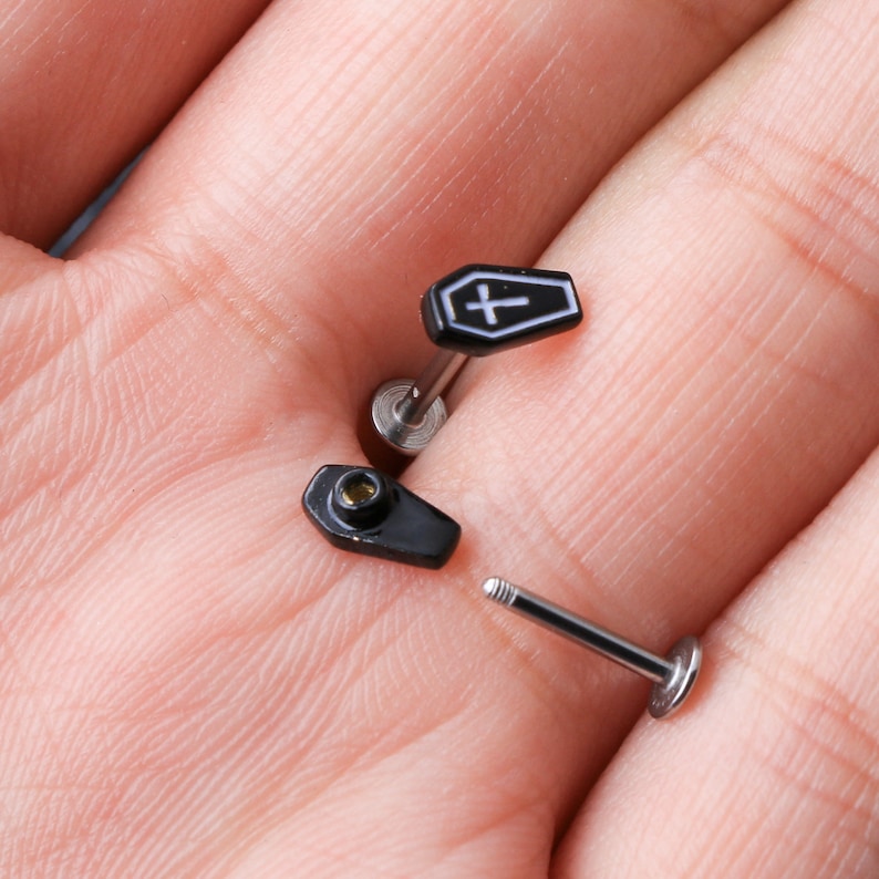 May include: Three black and silver coffin-shaped body jewelry pieces. Two of the pieces have a silver post and a black coffin with a white cross on top. The third piece is a silver post with a black coffin-shaped stud.