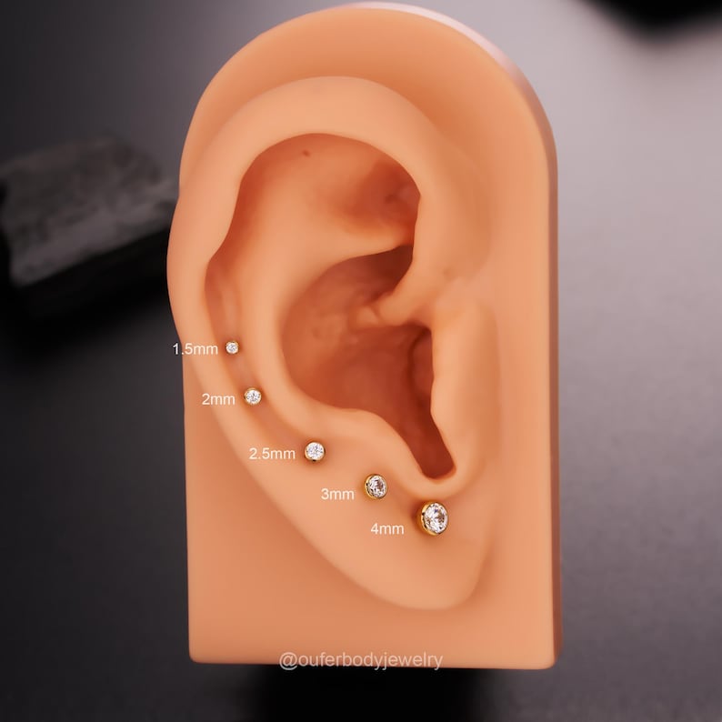 May include: A close-up of a model ear with five different sizes of clear gemstone earrings. The earrings are 1.5mm, 2mm, 2.5mm, 3mm, and 4mm in diameter. The text "@outerbodyjewelry" is at the bottom of the image.