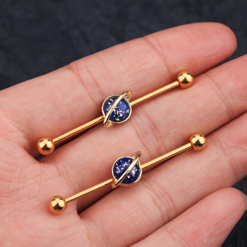 Puede incluir: Dos barras industriales doradas. Cada barra tiene un dije central azul en forma de planeta con un anillo dorado y extremos de bola dorados. Los dijes de planeta tienen un dise&ntilde;o moteado. Las joyas se muestran sobre un fondo neutro.