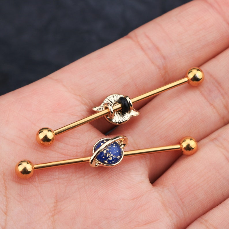 Puede incluir: Dos pendientes industriales dorados. Uno presenta un dise&ntilde;o de planeta azul con detalles dorados, y el otro tiene un dise&ntilde;o dorado. Cada pendiente tiene una bola en cada extremo.