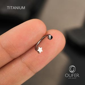 May include: A curved titanium body jewelry piece with a star and cone-shaped end. The word "TITANIUM" is printed in white in the upper left corner. The jewelry is silver in color and is held between two fingers.
