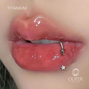 May include: Close-up of a person's lips with a lip piercing. The piercing is a silver-colored ring with a cone-shaped end. A small silver star is below the lip. The word "TITANIUM" is at the top left.
