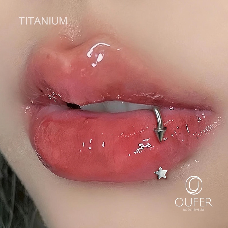 May include: Close-up of a person's lips with a lip piercing. The piercing is a silver-colored ring with a cone-shaped end. A small silver star is below the lip. The word "TITANIUM" is at the top left.