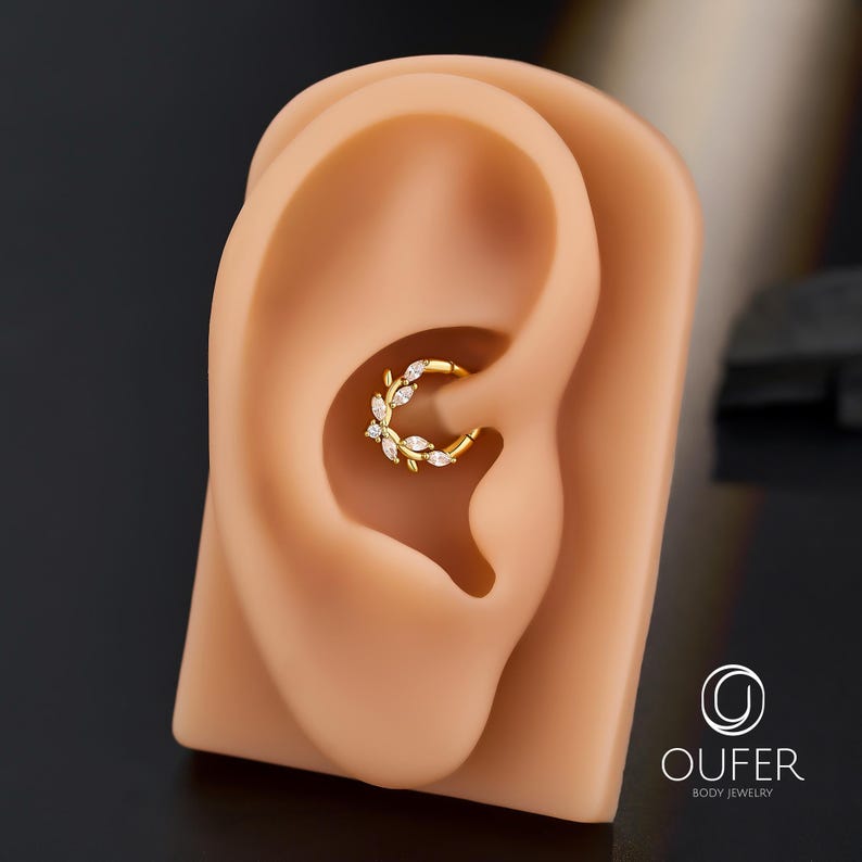 Op de afbeelding: Een goudkleurige oor piercing ring met een bladeren ontwerp, versierd met kleine, heldere edelstenen. De sieraden worden getoond op een tan oor model. De merknaam "OUFER BODY JEWELRY" is rechtsonder zichtbaar.