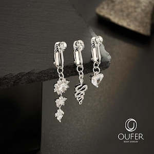 Puede incluir: Tres pendientes de clip en tono plateado con detalles de cristal transparente. El primer pendiente tiene un diseño floral colgante, el segundo presenta un dije de serpiente enroscada y el tercero muestra un colgante de cristal en forma de corazón. Logo OUFER.