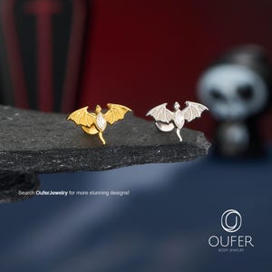 May include: Two bat-shaped stud earrings, one in gold tone and one in silver tone, each featuring a marquise-cut clear stone in the center. The text "Search OuferJewelry for more stunning designs!" is visible, along with the "OUFER BODY JEWELRY" logo.