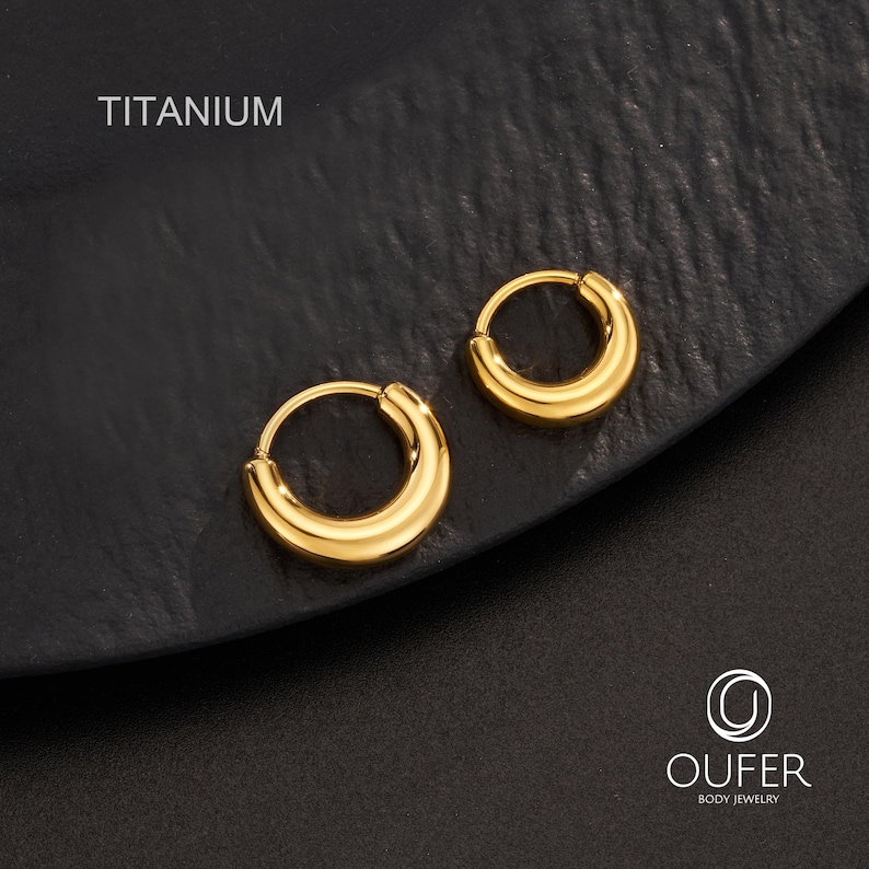 Puede incluir: Un par de pendientes de aro de titanio de color dorado. Los pendientes tienen forma de media luna y tienen un acabado liso y pulido. La palabra "TITANIUM" est&aacute; impresa en blanco en la parte superior de la imagen. Los pendientes se muestran sobre una superficie negra.