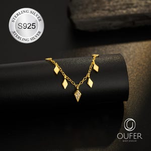 May include: A gold-colored necklace with diamond-shaped charms and a central diamond-shaped pendant. The necklace is displayed on a black surface. The image includes a silver emblem with the text "STERLING SILVER" and "S925". The brand name "OUFER BODY JEWELRY" is also visible.