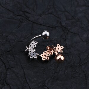 May include: Two silver and rose gold belly button rings with a flower design. The rings are on a black textured surface.