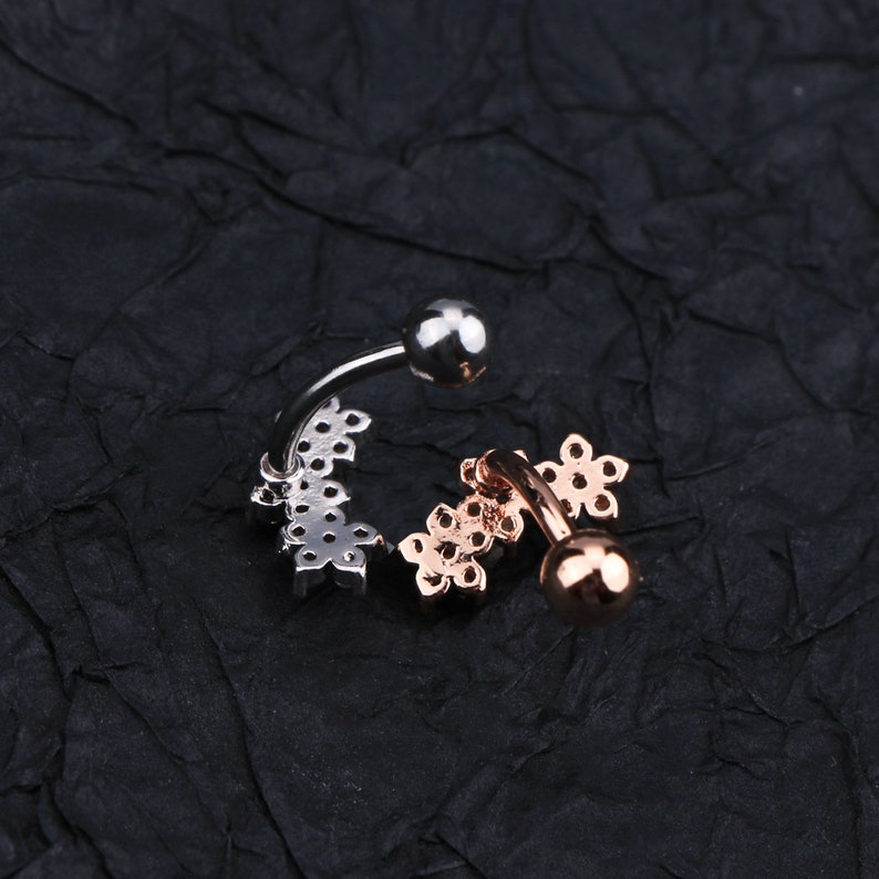 May include: Two silver and rose gold belly button rings with a flower design. The rings are on a black textured surface.