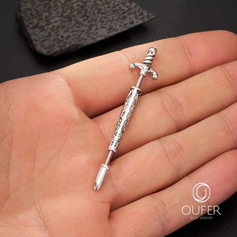 K&ouml;nnte beinhalten: Ein silberfarbenes, schwertf&ouml;rmiges K&ouml;rperschmuckst&uuml;ck. Der Schmuck hat ein detailliertes Griff- und Klingendesign. Der K&ouml;rperschmuck wird in der Hand einer Person gehalten.