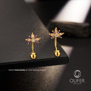 Può includere: Due gioielli per il corpo a barra curva color oro con una parte superiore a forma di fiore, decorata con pietre color pesca e viola taglio marquise attorno a una pietra centrale trasparente. Il testo recita: Search OuferJewelry for more stunning designs! OUFER BODY JEWELRY.