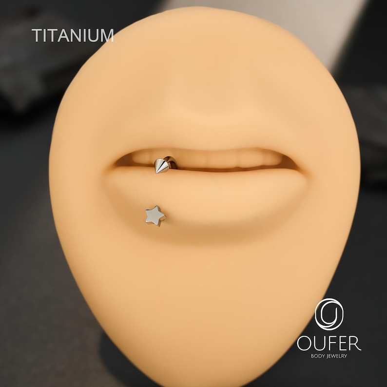 May include: A close-up of a mannequin's mouth with two pieces of body jewelry. One is a silver star-shaped stud, and the other is a silver cone-shaped lip ring. The word "TITANIUM" is printed in the upper left corner.