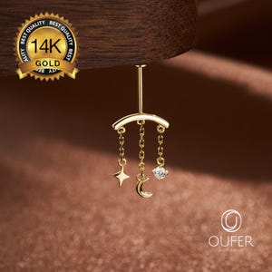 May include: A 14K gold belly button ring featuring a curved bar with three dangling chains. The chains hold a four-pointed star charm, a crescent moon charm, and a small round clear gemstone. A gold seal reads 14K GOLD BEST QUALITY.