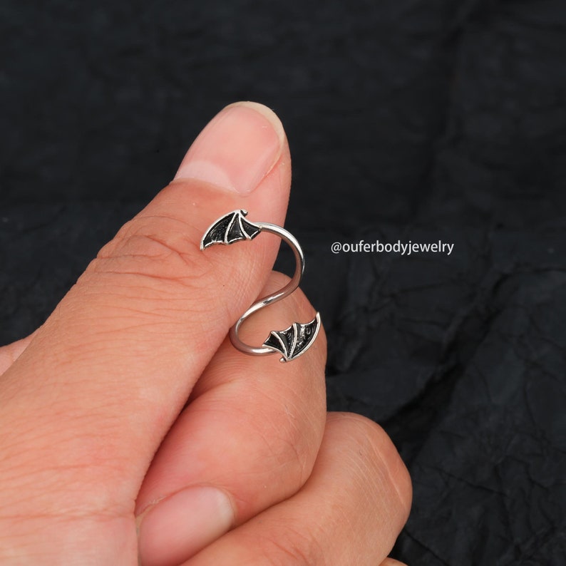Peut inclure: Piercing d'oreille en spirale argent&eacute; avec des ailes de chauve-souris noires &agrave; chaque extr&eacute;mit&eacute;. Le piercing est pr&eacute;sent&eacute; sur un doigt.