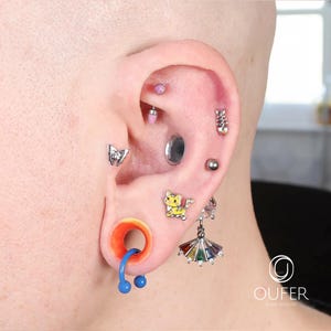 May include: An ear adorned with various body jewelry, including a silver butterfly stud, a small yellow cat stud, a silver fishbone stud, a silver ball stud, and a dangling umbrella earring with colorful gems. A large orange and blue ear gauge is also visible.