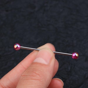 May include: A silver barbell with iridescent pink balls on each end. The barbell is held between two fingers.