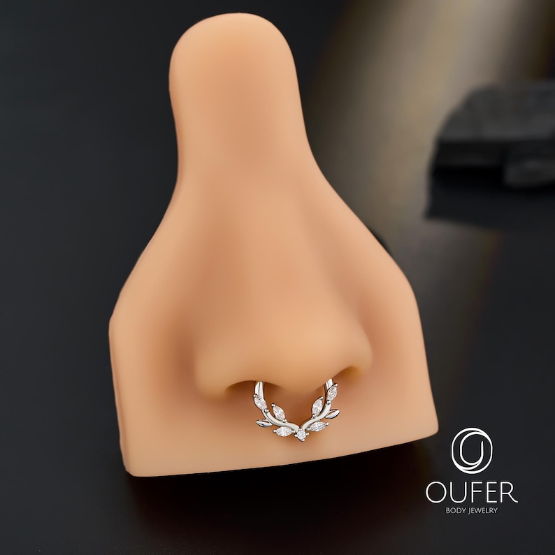 Op de afbeelding: Een zilveren septumring met een bladeren ontwerp, versierd met heldere edelstenen. De sieraden worden getoond op een neusmodel tegen een donkere achtergrond. Het merklogo "OUFER BODY JEWELRY" is rechtsonder zichtbaar.