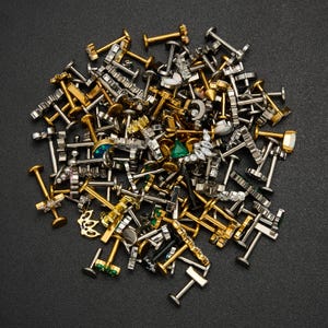 Puede incluir: Una colecci&oacute;n de piercings para labios y labret de varios dise&ntilde;os en tonos dorados y plateados, con diferentes adornos decorativos. Algunos piercings presentan piedras preciosas y dise&ntilde;os &uacute;nicos, incluyendo formas geom&eacute;tricas y motivos florales. Los piercings est&aacute;n dispersos sobre una superficie oscura.