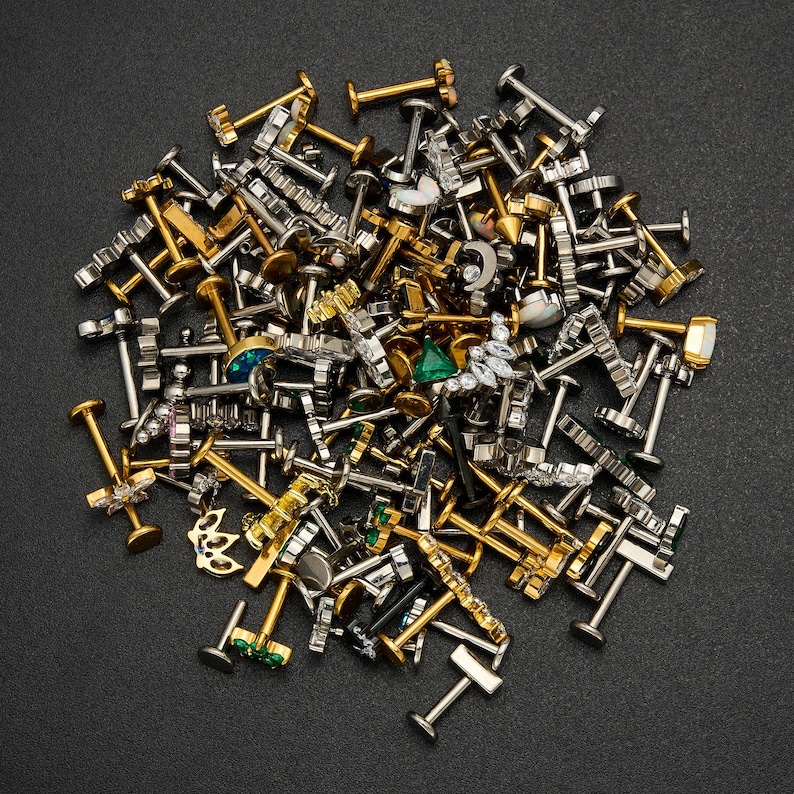 Puede incluir: Una colecci&oacute;n de piercings para labios y labret de varios dise&ntilde;os en tonos dorados y plateados, con diferentes adornos decorativos. Algunos piercings presentan piedras preciosas y dise&ntilde;os &uacute;nicos, incluyendo formas geom&eacute;tricas y motivos florales. Los piercings est&aacute;n dispersos sobre una superficie oscura.