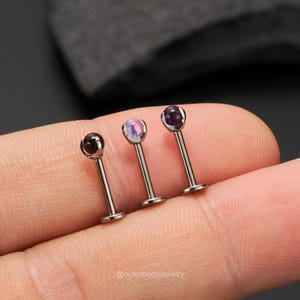 Peut inclure: Trois boucles d'oreilles barbell argentées avec des pierres précieuses de différentes couleurs. Les pierres précieuses sont l'opale, l'améthyste et l'onyx noir.
