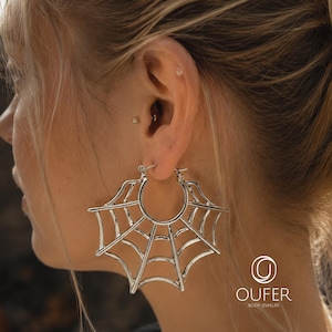 May include: A silver hoop earring featuring a prominent spider web design that extends from the circular band. The metal has a polished finish and the earring is secured through the earlobe. The logo OUFER BODY JEWELRY is visible in the corner.