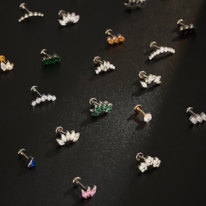 Puede incluir: Una variedad de pendientes de bot&oacute;n con varios dise&ntilde;os, incluyendo formas de hojas y racimos. Los pendientes presentan piedras preciosas claras, verdes, rosas y azules, engastadas en tonos dorados y plateados. Los pendientes se exhiben sobre una superficie oscura.
