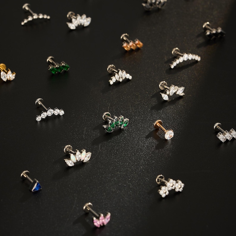Puede incluir: Una variedad de pendientes de bot&oacute;n con varios dise&ntilde;os, incluyendo formas de hojas y racimos. Los pendientes presentan piedras preciosas claras, verdes, rosas y azules, engastadas en tonos dorados y plateados. Los pendientes se exhiben sobre una superficie oscura.