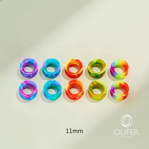 May include: A collection of ten colorful, rainbow-swirled silicone ear gauges. Each gauge is 11mm in diameter. The gauges are arranged in two rows of five, showcasing a variety of vibrant colors including purple, blue, red, yellow, and green. The brand name "OUFER BODY JEWELRY" is visible.