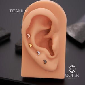 May include: Four crescent moon-shaped earrings in silver, gold, black, and light pink are displayed on a tan ear model. The word "TITANIUM" is printed in white at the top left. The earrings are body jewelry.