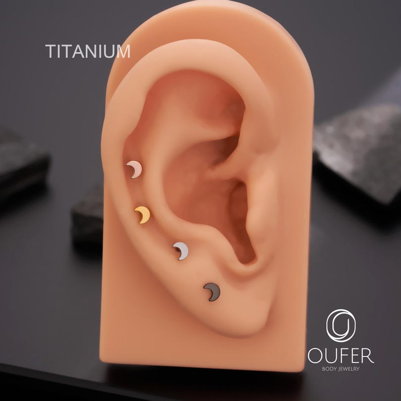 May include: Four crescent moon-shaped earrings in silver, gold, black, and light pink are displayed on a tan ear model. The word "TITANIUM" is printed in white at the top left. The earrings are body jewelry.