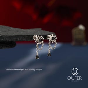 May include: A pair of silver-toned stud earrings featuring a spider web design with a central black stone. Each earring has a delicate silver chain dangling from the web, ending in a small, black, pear-shaped stone drop. Text reads: Search OuferJewelry for more stunning designs! OUFER BODY JEWELRY.