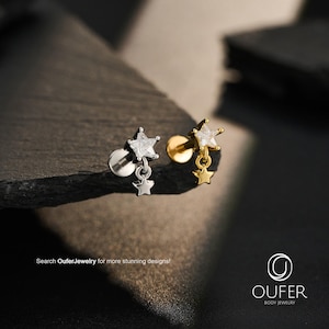 May include: Two flat back labret studs featuring a star-shaped clear crystal top with a small dangling star charm. One stud is silver-toned and the other is gold-toned. The text "Search OuferJewelry for more stunning designs!" and the Oufer logo are visible.