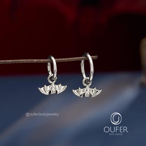 May include: A pair of silver-toned hoop earrings, each featuring a small, detailed bat charm hanging from the bottom. The text @ouferbodyjewelry and the logo OUFER BODY JEWELRY are visible in the lower portion of the image.