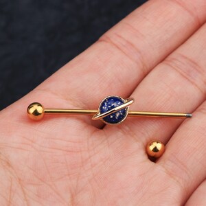 Puede incluir: Pendiente industrial de barra dorada con un dise&ntilde;o de planeta azul, un anillo dorado y motas blancas. El pendiente tiene una bola dorada en cada extremo. Tambi&eacute;n se ve una bola dorada separada.
