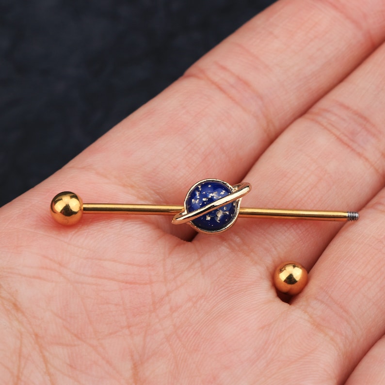 Puede incluir: Pendiente industrial de barra dorada con un dise&ntilde;o de planeta azul, un anillo dorado y motas blancas. El pendiente tiene una bola dorada en cada extremo. Tambi&eacute;n se ve una bola dorada separada.