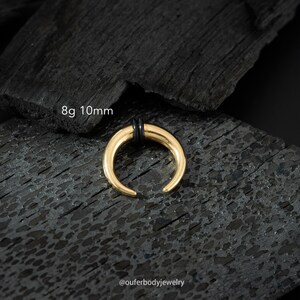 May include: Gold 8g 10mm horn septum ring with black o-rings. The ring is on a black background.