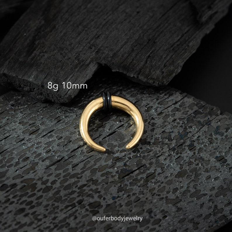 May include: Gold 8g 10mm horn septum ring with black o-rings. The ring is on a black background.