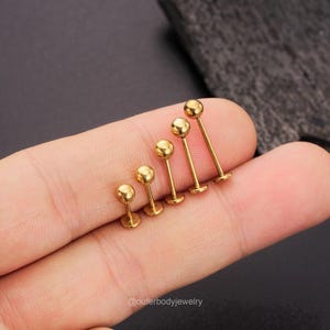 May include: A set of six gold-colored barbell earrings with different sized balls. The earrings are arranged in order of size, with the smallest on the left and the largest on the right. The earrings are held in a hand.