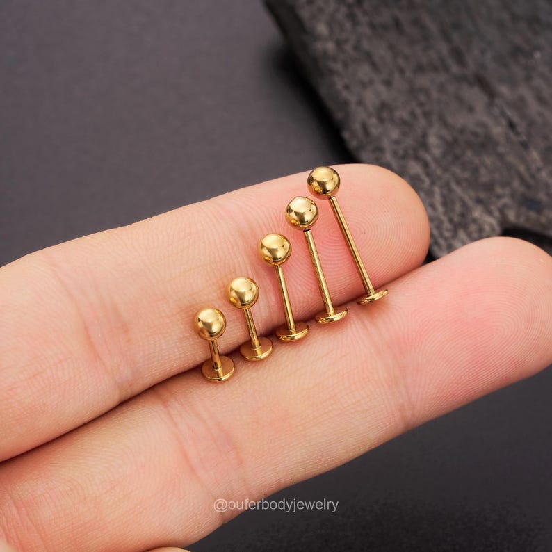 May include: A set of six gold-colored barbell earrings with different sized balls. The earrings are arranged in order of size, with the smallest on the left and the largest on the right. The earrings are held in a hand.
