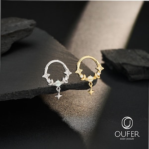 May include: Two circular hoop rings, one silver-toned and one gold-toned, each featuring a central opal-like stone, small crescent moons, star-shaped accents with embedded clear crystals, and a dangling star charm. The text OUFER BODY JEWELRY is visible.